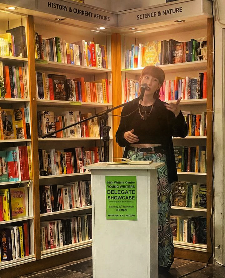 HK is standing in front of a shelf of books performing a poem. 