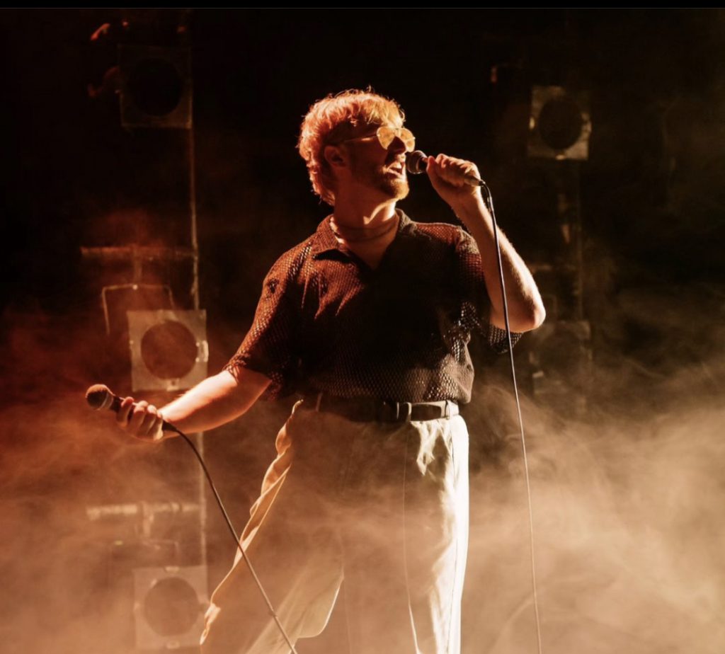 A man singing into a microphone in a black top and white pants. He is surrounded by an orange mist