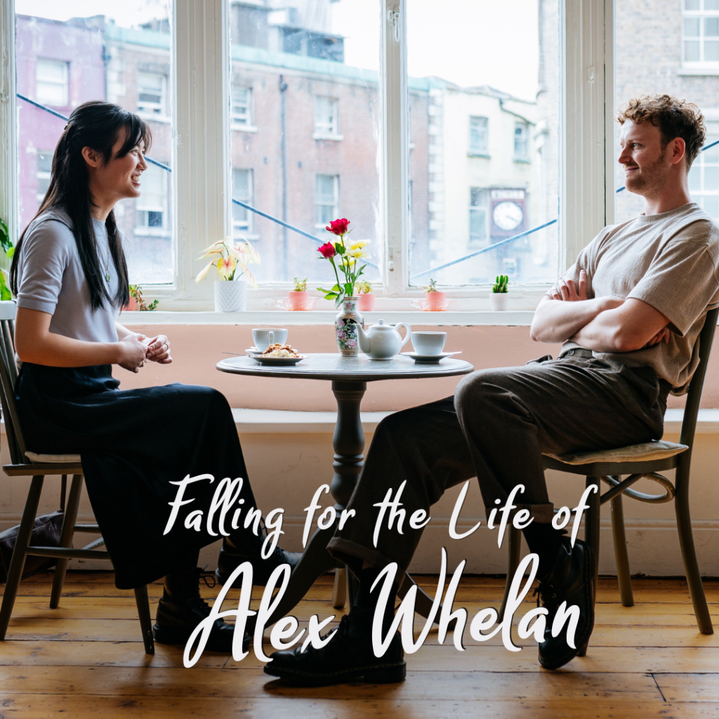 An asian woman sitting across from a white man at a café. They are sitting in front of a window which is ornate. Above them is the text 'Falling for the Life of Alex Whelan'
